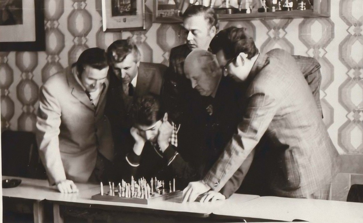 Sechs Männer, meist in Anzügen, stehen dicht gedrängt um ein Schachbrett auf einem Tisch und beobachten aufmerksam das Spiel. Der Schauplatz scheint ein Innenraum mit gemusterten Tapeten und Regalen im Hintergrund zu sein.