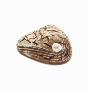 Ein dreieckiger Laib rustikales Schwarzbrot mit Mehl bestäubt und einem runden weißen Etikett auf der Oberseite, das auf einem weißen Hintergrund ruht. Das Brot hat eine rissige, strukturierte Kruste mit dekorativen Einkerbungen.