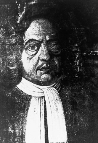 Schwarz-Weiß-Porträt eines Mannes mit lockigem Haar, markanten Augen und ernstem Gesichtsausdruck, der einen dunklen Mantel und ein weißes Halstuch trägt. Der Hintergrund ist dunkel und teilweise unscharf.