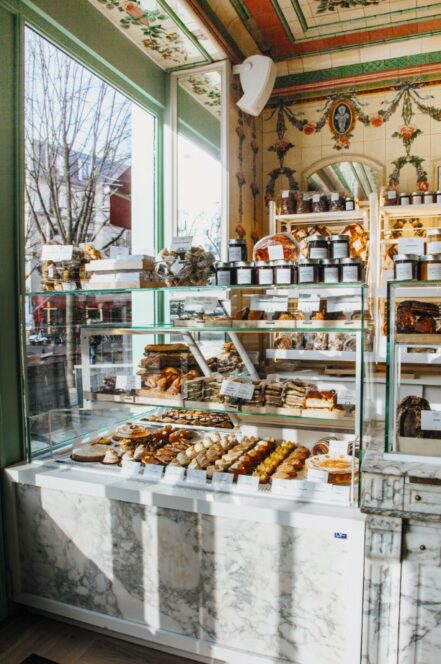 In einer hellen, eleganten Bäckerei mit Marmortheken und kunstvollem Dekor sind die Regale mit verschiedenen Backwaren, Broten und Gläsern gefüllt. Das Sonnenlicht strömt durch die großen Fenster und bringt die Backwaren zur Geltung.