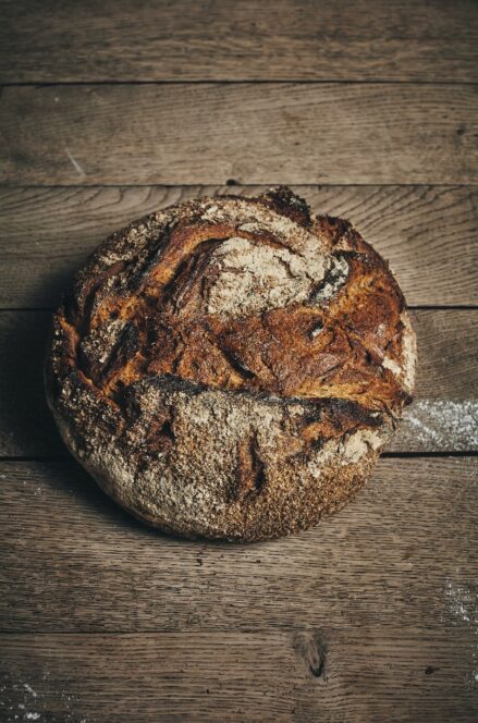Ein runder, rustikaler Laib Sauerteigbrot mit einer dunklen, knusprigen Oberfläche liegt auf einer mit Mehl bestäubten Holzfläche.
