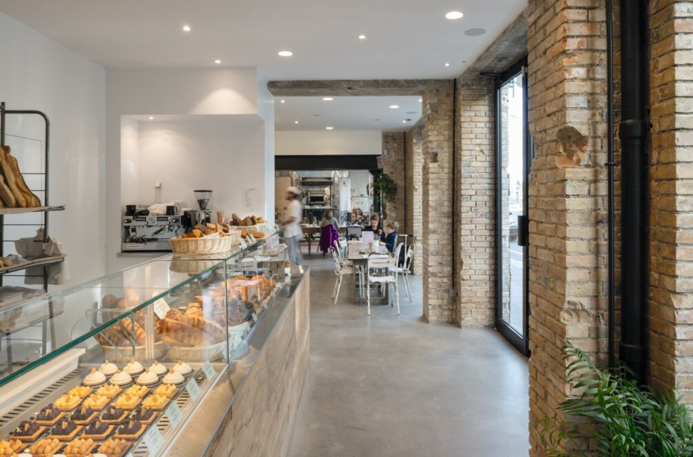 Eine moderne Bäckerei mit freiliegenden Backsteinwänden, einer Glasvitrine mit Gebäck, Brot in Regalen und einem Sitzbereich mit Tischen und Stühlen. Große Fenster lassen viel natürliches Licht herein.