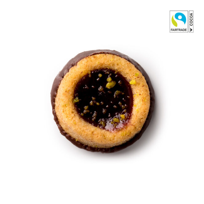 Ein runder Keks, halb in Schokolade getaucht, gefüllt mit roter Marmelade und gekrönt mit kleinen grünen Nussstückchen, auf weißem Hintergrund mit einem Fairtrade-Kakao-Etikett in der oberen rechten Ecke.
