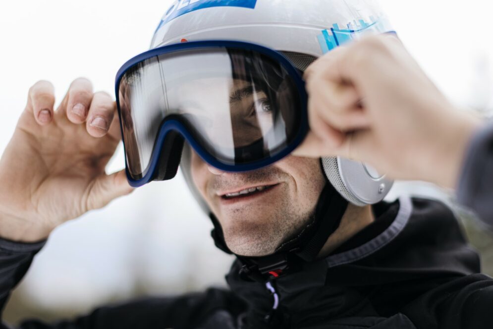 Eine Person, die einen Helm trägt, ihre Skibrille zurechtrückt und leicht lächelt. Das Foto ist im Freien aufgenommen, möglicherweise in einem Skigebiet oder an einem verschneiten Ort.