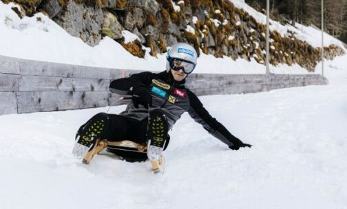 Eine Person in Wintersportkleidung und mit Helm fährt auf einem Holzschlitten eine verschneite Piste hinunter und steuert mit einer Hand auf dem Schnee. Am Rande der Strecke verläuft eine felsige, schneebedeckte Böschung.