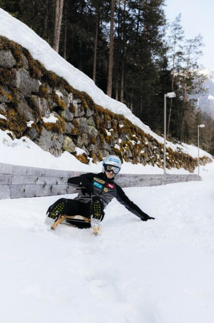 Eine Person in Winterausrüstung und mit Helm gleitet auf einem Holzschlitten einen verschneiten Hang hinunter, wobei eine Hand den Schnee berührt. Kiefern und eine Steinmauer säumen den Hintergrund.