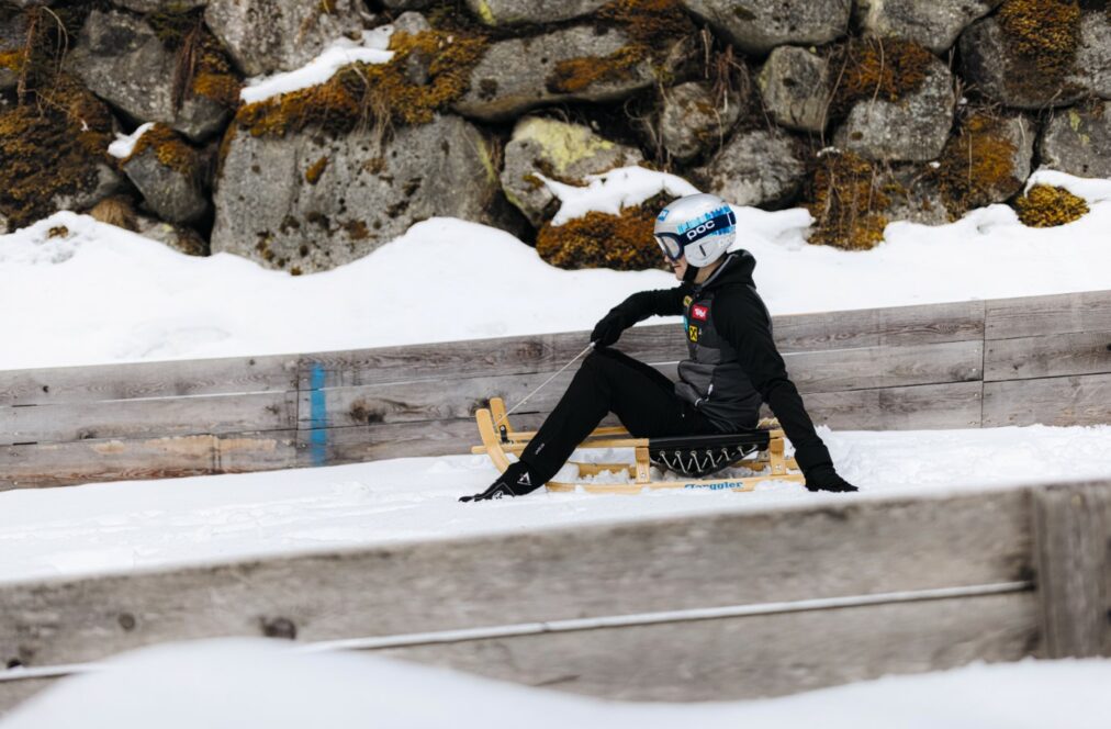 Eine Person in Winterausrüstung und mit Helm sitzt auf einem kleinen Holzschlitten auf einem verschneiten Weg neben einer mit Moos bewachsenen Steinmauer.