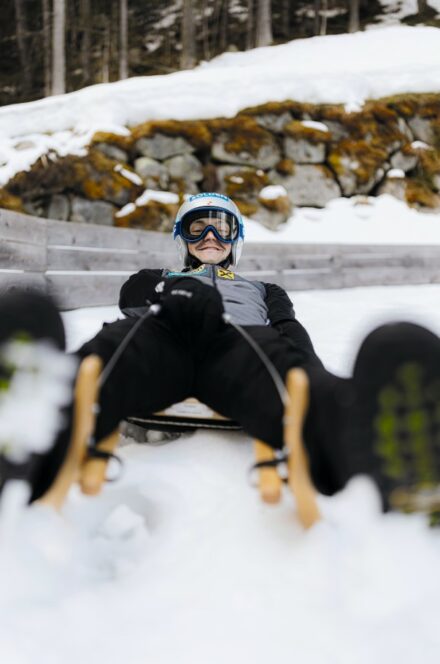 Eine Person, die einen Helm, eine Schutzbrille und Winterkleidung trägt, fährt auf einem Holzschlitten einen verschneiten Abhang hinunter, mit Bäumen, Felsen und einer Holzbarriere im Hintergrund. Das Foto ist aus einem niedrigen Winkel vor dem Schlitten aufgenommen.