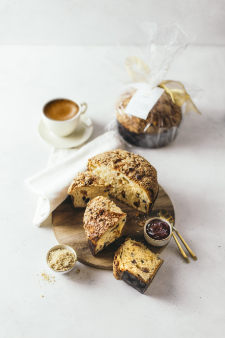 Ein runder, in Scheiben geschnittener Laib süßes Brot auf einem Holzbrett, dazu eine Tasse Espresso, ein Glas Marmelade, eine kleine Schale mit Zucker und goldenes Besteck. Ein weiteres eingewickeltes Brot liegt im Hintergrund.