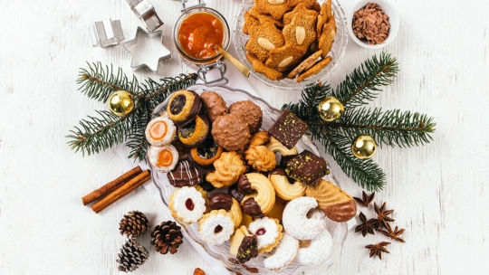 Ein festliches Arrangement aus verschiedenen Weihnachtsplätzchen und Pralinen auf einer Glasplatte, umgeben von Tannenzweigen, goldenen Ornamenten, Zimtstangen und Sternanis auf einem weißen Holztisch.