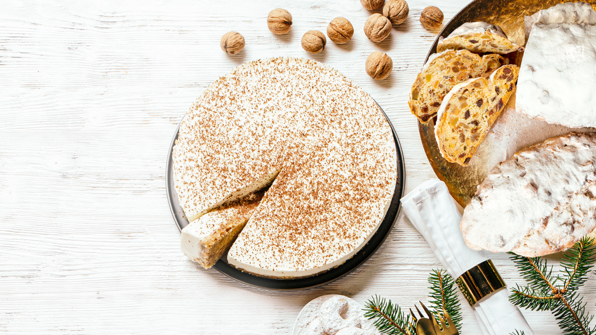 Ein runder Käsekuchen, leicht mit Zimt bestäubt, von dem ein Stück abgeschnitten wurde. Daneben liegen Walnüsse, geschnittenes Brot, mit Puderzucker bestäubtes Gebäck, eine Serviette mit goldenem Besteck und Tannenzweige – alles auf einem weißen Holztisch.