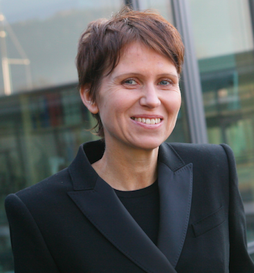 Eine Person mit kurzen braunen Haaren, die einen schwarzen Blazer trägt und lächelt, steht im Freien vor einer Glasscheibe.