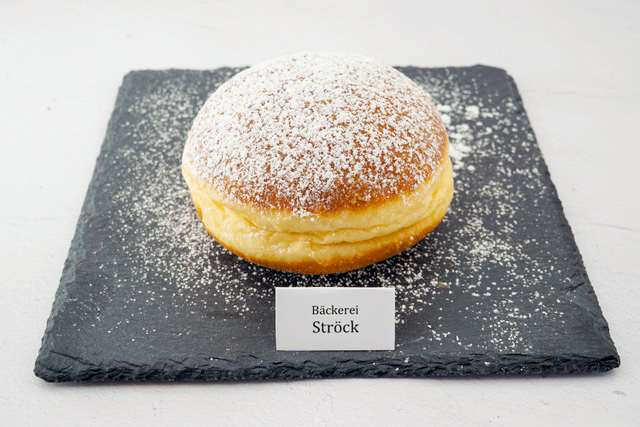 Ein rundes, mit Puderzucker bestreutes Gebäck liegt auf einer dunklen Schieferplatte, vor der ein kleines Schild mit der Aufschrift Bäckerei Ströck steht. Der Hintergrund ist eine schlichte, helle Fläche.