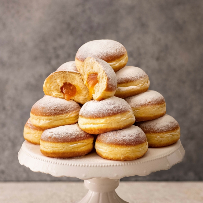 Ein Stapel mit Puderzucker gefüllter Donuts ist auf einem weißen Kuchenständer angeordnet, wobei ein Donut aufgeschnitten ist und eine karamellartige Füllung zum Vorschein kommt. Der Hintergrund ist ein neutrales Grau.