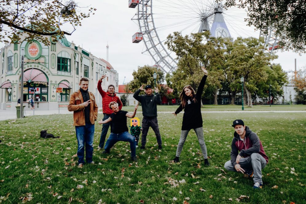 Sechs Personen posieren spielerisch im Gras vor einem Riesenrad und einem historischen Gebäude, umgeben von Bäumen und heruntergefallenen Blättern. Einige stehen, einige hocken, alle lächeln und machen lebhafte Gesten.