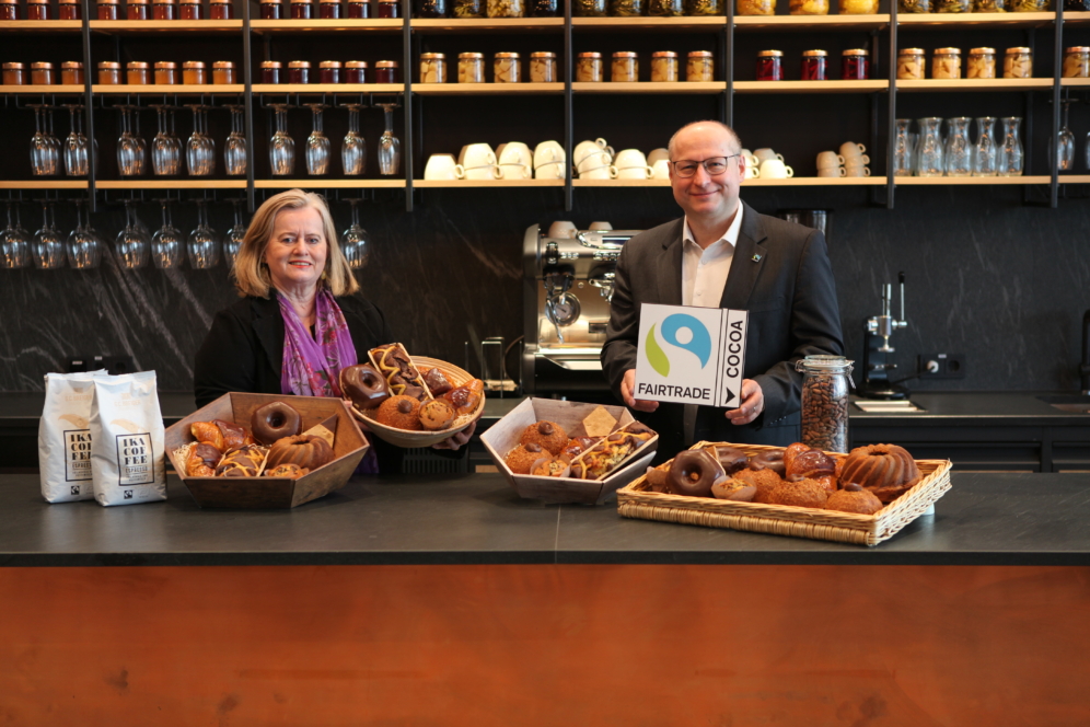 Zwei Personen stehen hinter einer Theke mit Körben voller Gebäck und Donuts. Der Mann hält ein Schild mit Fairtrade-Kakao in der Hand. Im Hintergrund stehen Regale mit Gläsern und Tassen, und auf dem Tresen stehen Tüten mit Kaffee.