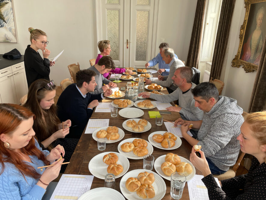 Eine Gruppe von Menschen sitzt um einen langen Tisch mit Brötchentellern, Wassergläsern und Bewertungsbögen und scheint an einer Verkostung oder Bewertung in einem hellen Raum mit Gemälden an den Wänden teilzunehmen.