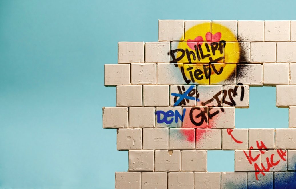 Eine weiße Backsteinmauer mit einem Loch, bedeckt mit bunten Graffiti, darunter ein gelbes Smiley-Gesicht mit einem Herz und verschiedene deutsche Wörter und Tags. Der Hintergrund ist einfarbig hellblau.