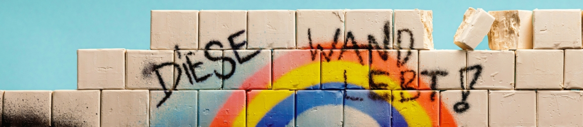 Weiße Blöcke bilden eine Wand mit einem Regenbogen und schwarzem Graffiti-Text in Deutsch mit der Aufschrift DIESE WAND BLEIBT? vor blauem Hintergrund; ein Block an der Spitze scheint zu fallen.