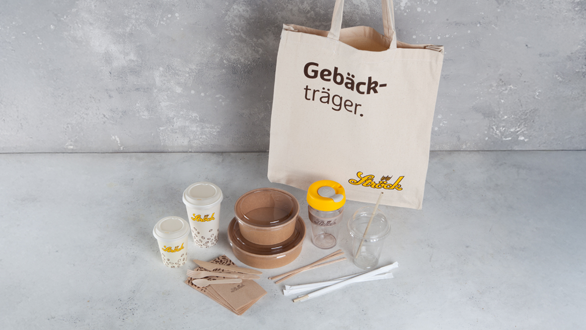 Eine beigefarbene Tragetasche mit dem Aufdruck Gebäckträger ist umgeben von verschiedenen umweltfreundlichen Mitnahmeartikeln wie Bechern, Schüsseln, Gläsern, Besteck, Servietten und Strohhalmen auf einer hellgrauen Fläche.