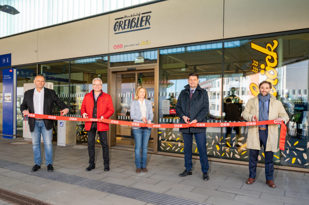 Fünf Personen stehen vor einem Gebäude und halten ein rotes Band für eine Zeremonie zum Durchschneiden des Bandes. Das Gebäude hat Glastüren und Schilder mit den Aufschriften GREISBLER und OBB über dem Eingang.