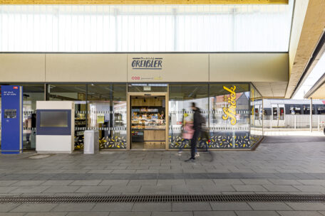 Ein Bäckereifachgeschäft an einem Bahnhof mit Glaswänden, die Backwaren ausstellen. Zwei verschwommene Personen gehen vorbei, und rechts ist ein Zug zu sehen. Auf dem Schild der Bäckerei steht "Greissler".