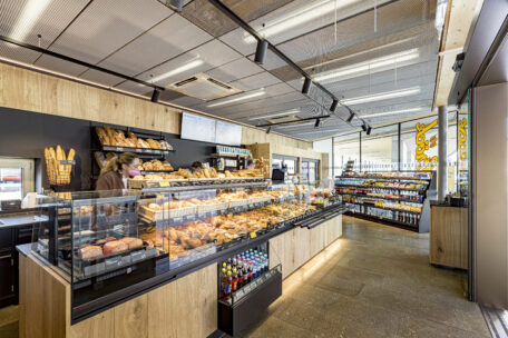 Eine moderne Bäckerei mit Holzregalen und Glasvitrinen, gefüllt mit verschiedenen Brotsorten und Gebäck. Hinter der Theke werden Kunden bedient, und im Hintergrund sind verpackte Waren in Regalen zu sehen.