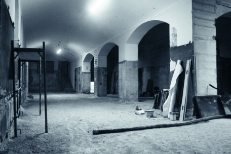 Ein großer, leerer, schwach beleuchteter Raum, der gerade renoviert wird, mit gewölbten Decken, freiliegenden Wänden, verstreuten Baumaterialien und einem Treppenhaus im Hintergrund.