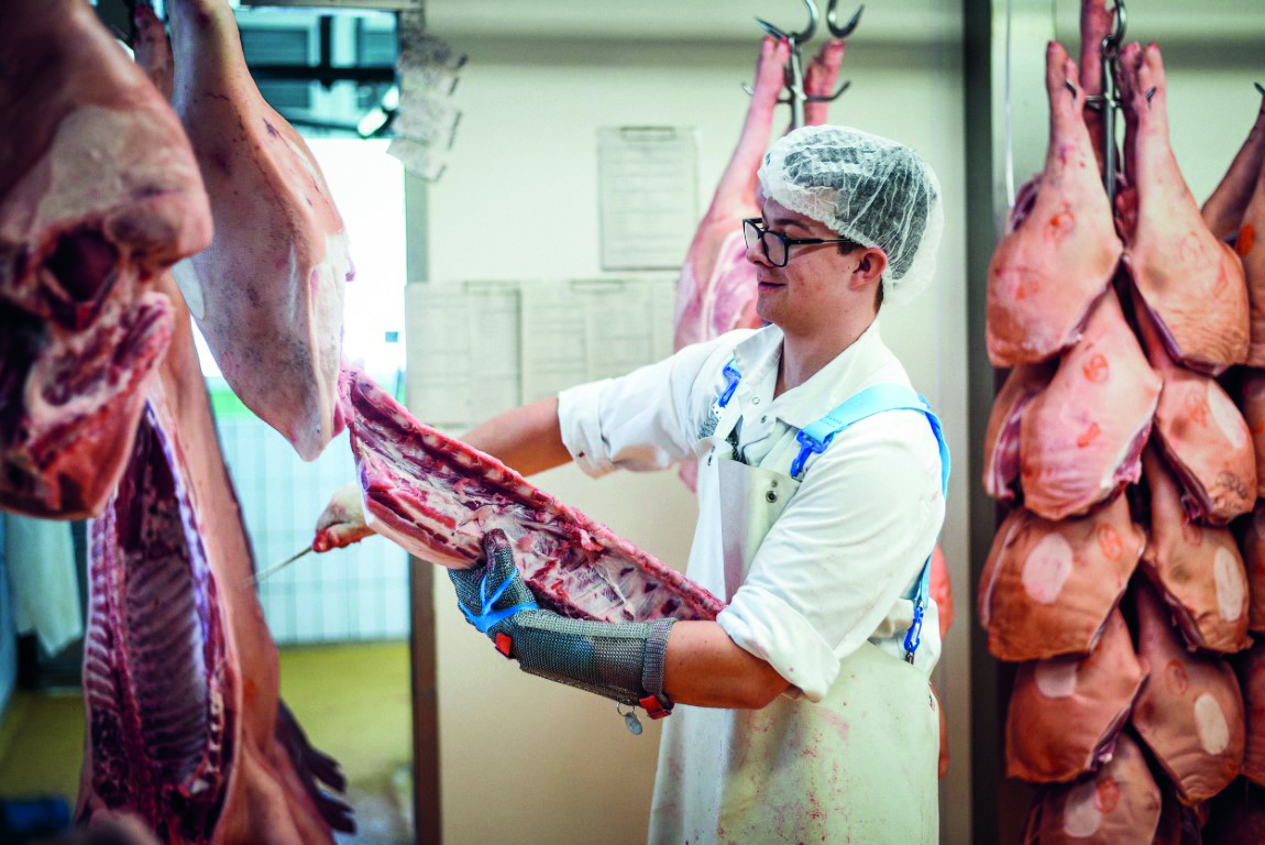 Ein Metzger, der Schutzkleidung und Brille trägt, schneidet in einem Fleischverarbeitungsbetrieb Fleisch von einem hängenden Schlachtkörper ab, umgeben von mehreren anderen hängenden Fleischstücken.