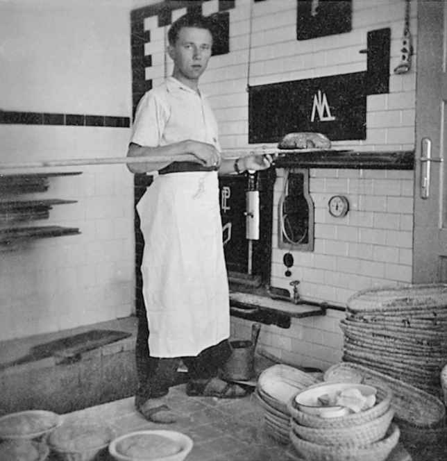 Ein Bäcker in weißer Schürze steht an einem Ofen und hält eine lange hölzerne Schale mit einem Laib Brot in der Hand. In einer gekachelten Bäckerei stehen Körbe und Schüsseln mit Teig auf dem Boden.