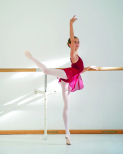Eine junge Ballerina in einem burgunderroten Trikot und einer weißen Strumpfhose führt an einer Ballettstange in einem hellen Studio im Sonnenlicht eine Arabeske vor.