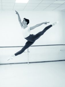 Ein Balletttänzer in der Luft führt in einem Tanzstudio einen Grand Jeté aus. Er trägt ein weißes Hemd, eine dunkle Strumpfhose und Spitzenschuhe. Ein Arm ist angehoben, und beide Beine sind gespreizt. Der Hintergrund ist hell und minimalistisch.