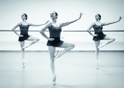 Drei Balletttänzerinnen in passenden Trikots und Röcken stehen auf einem Bein, die Arme anmutig ausgestreckt, und tanzen in einem Studio mit einer Barre im Hintergrund. Das Bild ist in schwarz-weiß gehalten.
