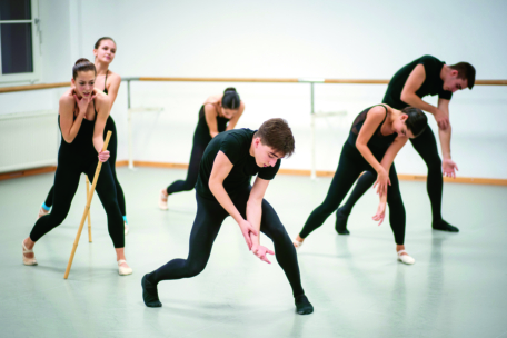 Sechs Balletttänzerinnen und -tänzer in schwarzen Kostümen zeigen ausdrucksstarke, dramatische Posen in einem Tanzstudio mit Spiegeln und einer Ballettstange. Eine Tänzerin hält einen Holzstab, während sich die anderen in verschiedenen Positionen beugen und strecken.