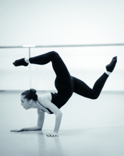 Eine Tänzerin in einem schwarzen Outfit balanciert auf einer Hand mit angewinkelten und in die Luft gehobenen Beinen und demonstriert so Kraft und Flexibilität in einem Tanzstudio mit einem Barre im Hintergrund.
