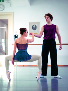 Eine Balletttänzerin in einer tiefen Plié-Pose auf der Spitze hält die Hand eines vor ihr stehenden männlichen Tänzers. Sie proben in einem Ballettstudio mit Spiegeln und einer Barre im Hintergrund.