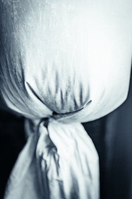 Nahaufnahme eines weißen, verknoteten Tuches, in dem Falten und Knicke zu sehen sind. Die Beleuchtung erzeugt einen weichen, fast monochromen Effekt, der die Textur des Stoffes und die sanften Schatten betont.