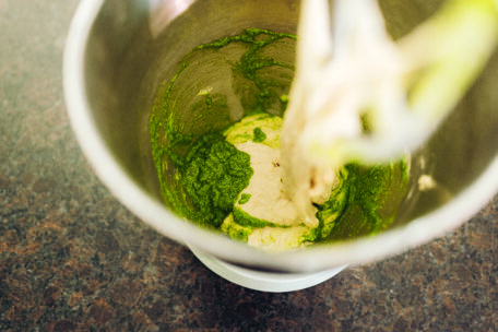 Eine Rührschüssel mit grüner Pesto-Soße und Teig darin, von oben auf einer dunklen Arbeitsplatte gezeigt, wobei ein Standmixer-Aufsatz teilweise sichtbar ist.