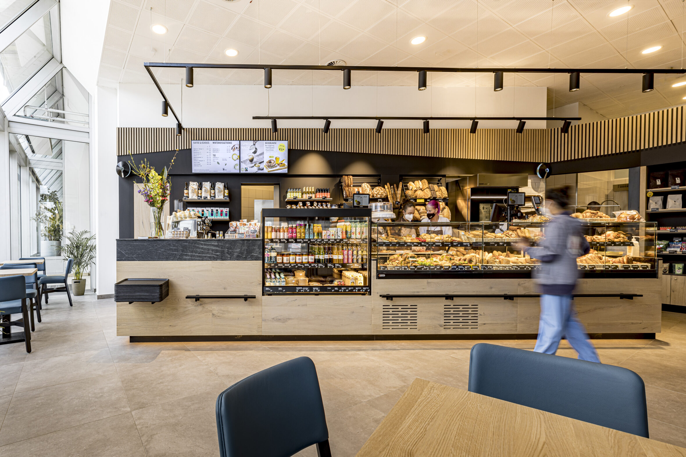 Ein modernes Bäckerei-Café mit Holztheken, auf denen Gebäck, Brot und Getränke angeboten werden. Über der Theke hängen Menütafeln, und in der Nähe steht eine Person in Bewegung. Blaue Stühle und Tische stehen im Vordergrund.