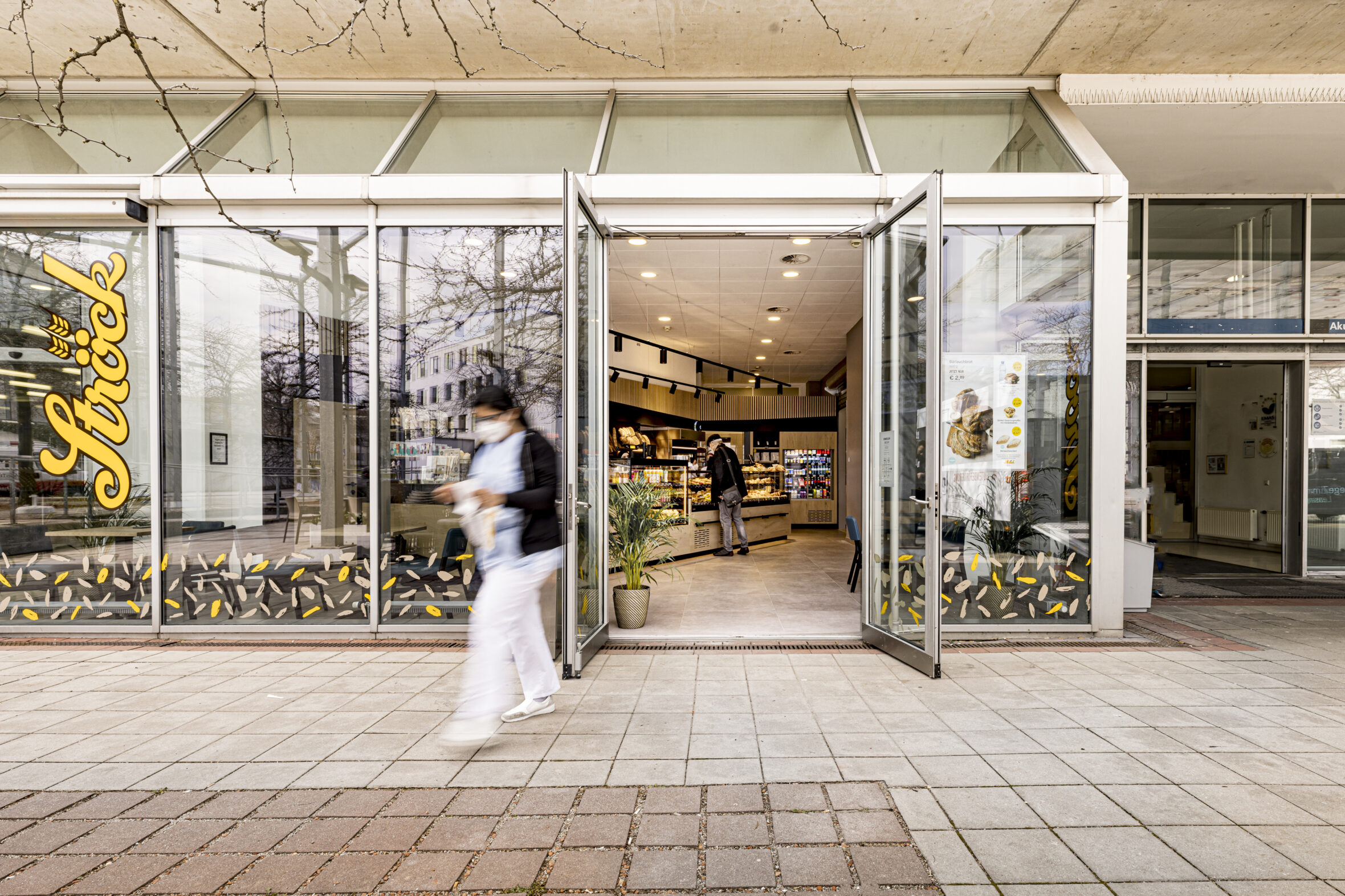 Eine Person verlässt eine moderne Bäckerei mit großen Glasfenstern und offenen Flügeltüren; eine andere Person steht drinnen in der Nähe der Theke. Die Bäckerei bietet Gebäck und Getränke an, und die Straße ist mit Fliesen gepflastert.