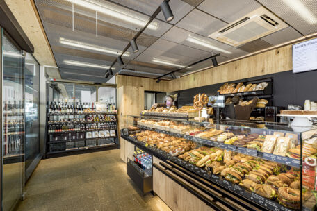 In einer modernen Bäckerei mit hölzernen Akzenten werden hinter einer Glastheke verschiedene Brotsorten, Gebäck und belegte Brötchen angeboten. Hinter der Theke steht ein Mitarbeiter mit einer Maske. Im Hintergrund befinden sich Regale mit Weinflaschen.