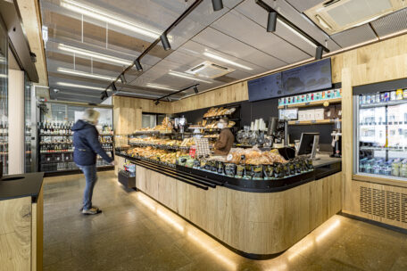 Eine moderne Bäckerei mit Holzdekor, mit Gebäck gefüllten Vitrinen, einer Mitarbeiterin hinter dem Tresen und einem Kunden, der sich umsieht. Im Hintergrund sind Regale mit Getränken zu sehen.