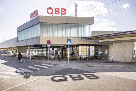 Ein modernes ÖBB-Bahnhofsgebäude mit großen Glasfenstern und einem Café im Inneren. Das ÖBB-Logo ist auf dem Dach zu sehen, und sein Schatten wird auf den Bürgersteig geworfen. Eine Person geht draußen in der Nähe eines Zebrastreifens.