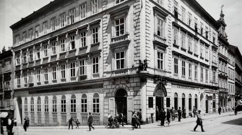 Schwarz-Weiß-Foto eines großen, historischen Eckgebäudes mit verschnörkelten architektonischen Details. Menschen gehen auf den Bürgersteigen und versammeln sich in der Nähe der Eingänge; die Straße ist belebt mit Fußgängern.