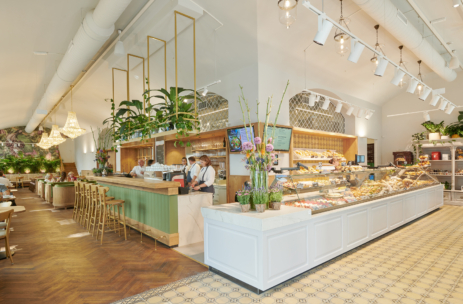 Helles, stilvolles Bäckerei-Café mit einer langen Vitrine voller Backwaren, mehreren Mitarbeitern hinter der Theke, grün-goldenem Dekor, Zimmerpflanzen, Kronleuchtern und gemütlichen Sitzecken mit Holzböden und gemusterten Fliesen.