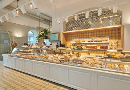 Eine helle Bäckerei mit einer langen Theke, die mit verschiedenen Brotsorten, Gebäck und Desserts gefüllt ist. Zwei Mitarbeiter stehen hinter der Theke, und der Raum ist mit gemusterten Bodenfliesen und warmer Beleuchtung ausgestattet.