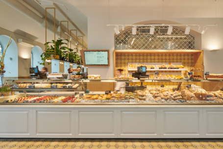 Eine helle, moderne Bäckerei mit einer großen Auslage, die mit verschiedenen Backwaren, Broten und Desserts gefüllt ist. Hinter der Theke sind die Regale mit Backwaren bestückt und an der Wand ist eine digitale Speisekarte zu sehen.