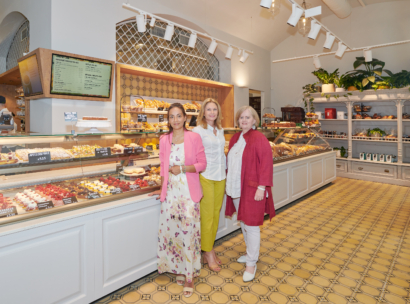 Drei Frauen stehen vor der gut beleuchteten Auslage einer Bäckerei, die mit Gebäck und Desserts gefüllt ist. Die Bäckerei hat geflieste Böden, Regale mit Pflanzen und Produkten und eine Tafel mit der Speisekarte über der Theke.