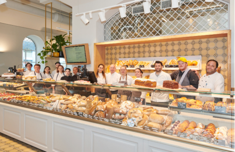 Eine Gruppe von Bäckereimitarbeitern steht hinter einer Glasvitrine mit verschiedenen Brotsorten, Gebäck und Kuchen in einer hellen, modernen Bäckerei mit gekachelten Wänden und Regalen mit Backwaren.