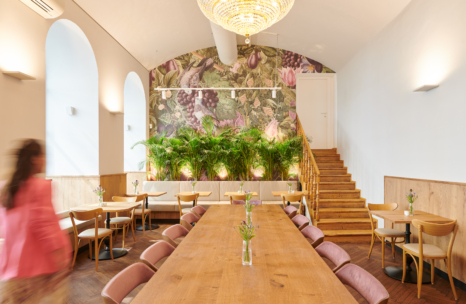 Ein geräumiges, helles Café mit einem langen Holztisch, rosafarbenen Stühlen, kleinen Tischen mit Holzstühlen, üppigen Grünpflanzen, Blumentapeten, Bogenfenstern, einem großen Kronleuchter und einer vorbeigehenden Person.